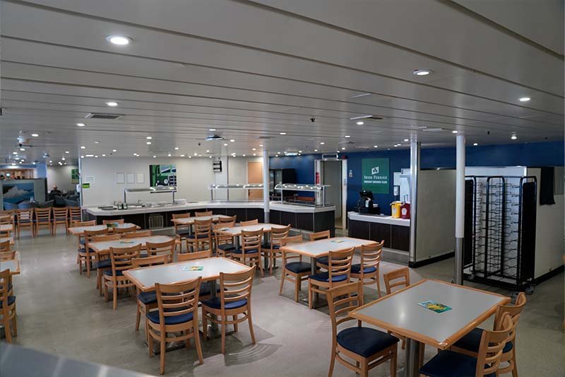 Self-service d’Irish Ferries avec des chaises en bois vides aux assises bleues, des tables blanches et une ligne de buffet au fond