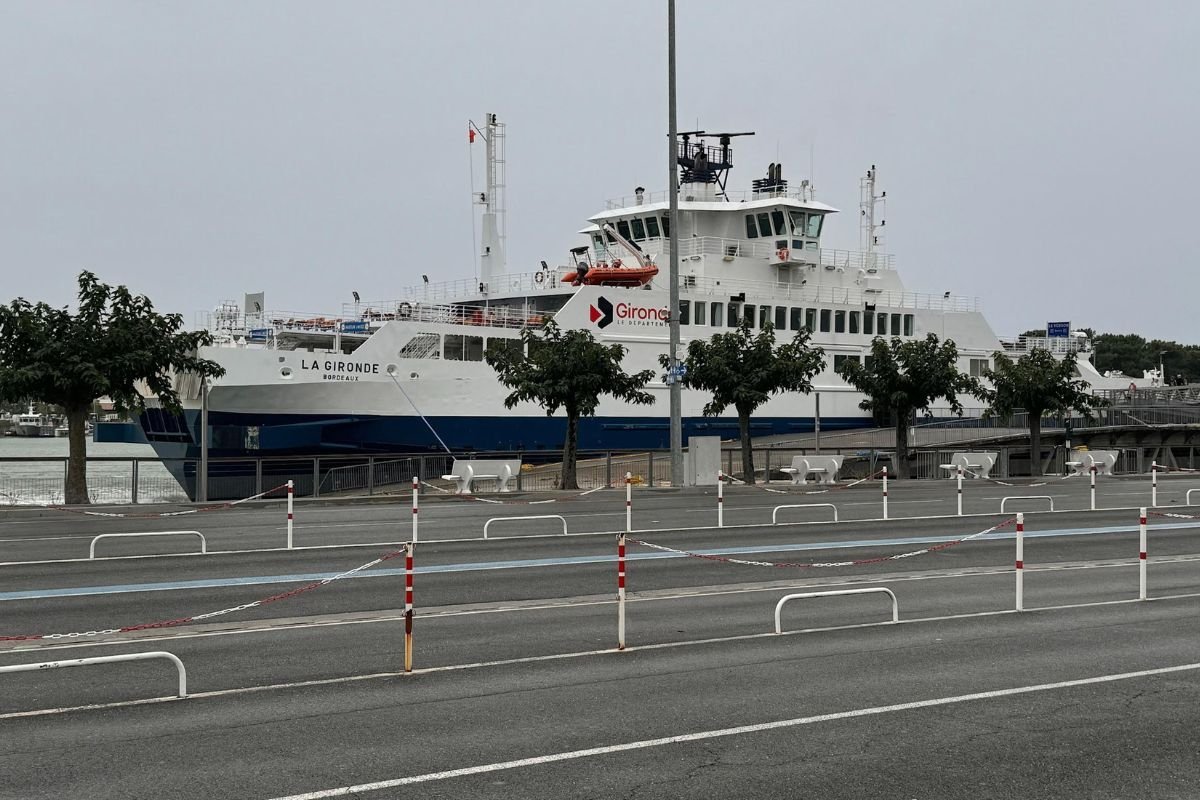 Le bac blanc La Gironde, de Bordeaux, à quai, vu au-delà de files d’attente vides bordées de plots rouges et blancs et de chaînes, sous un ciel gris