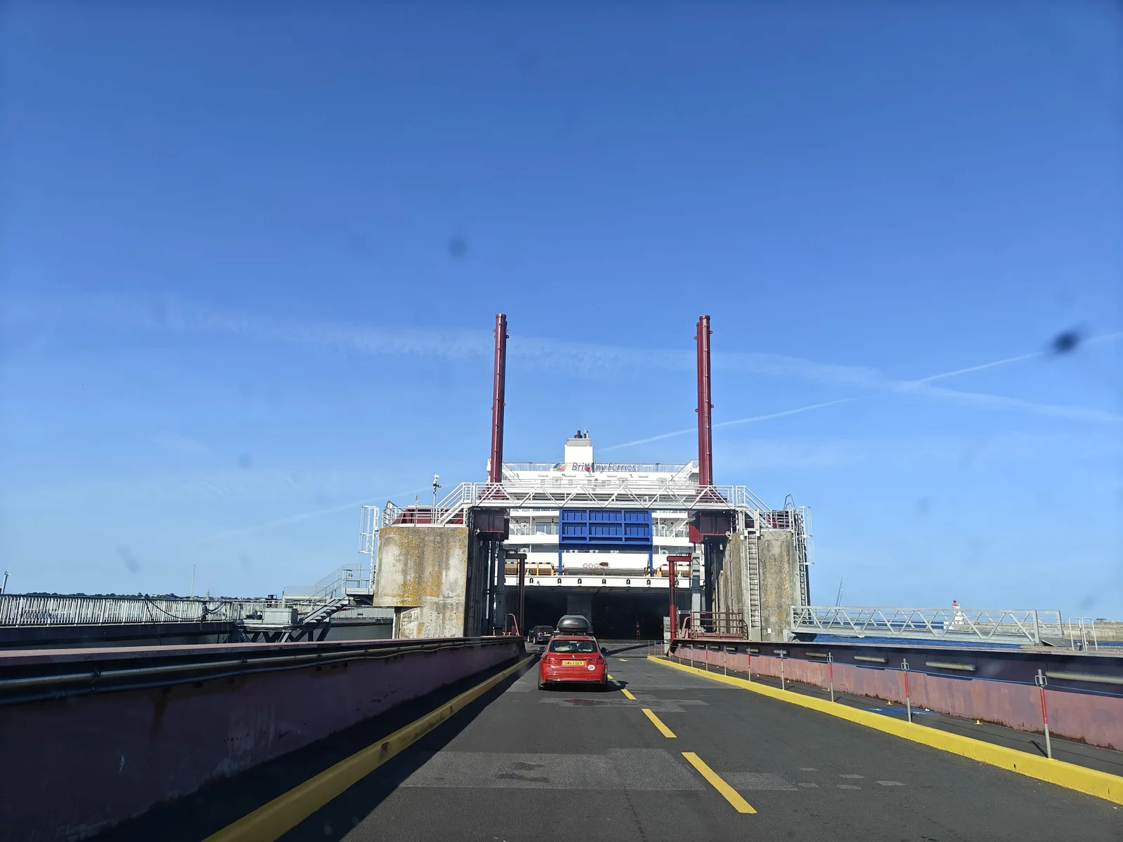 Une voiture rouge monte par la rampe dans la proue ouverte d’un ferry blanc de Brittany Ferries, sous un ciel bleu, dans le port de Saint-Malo