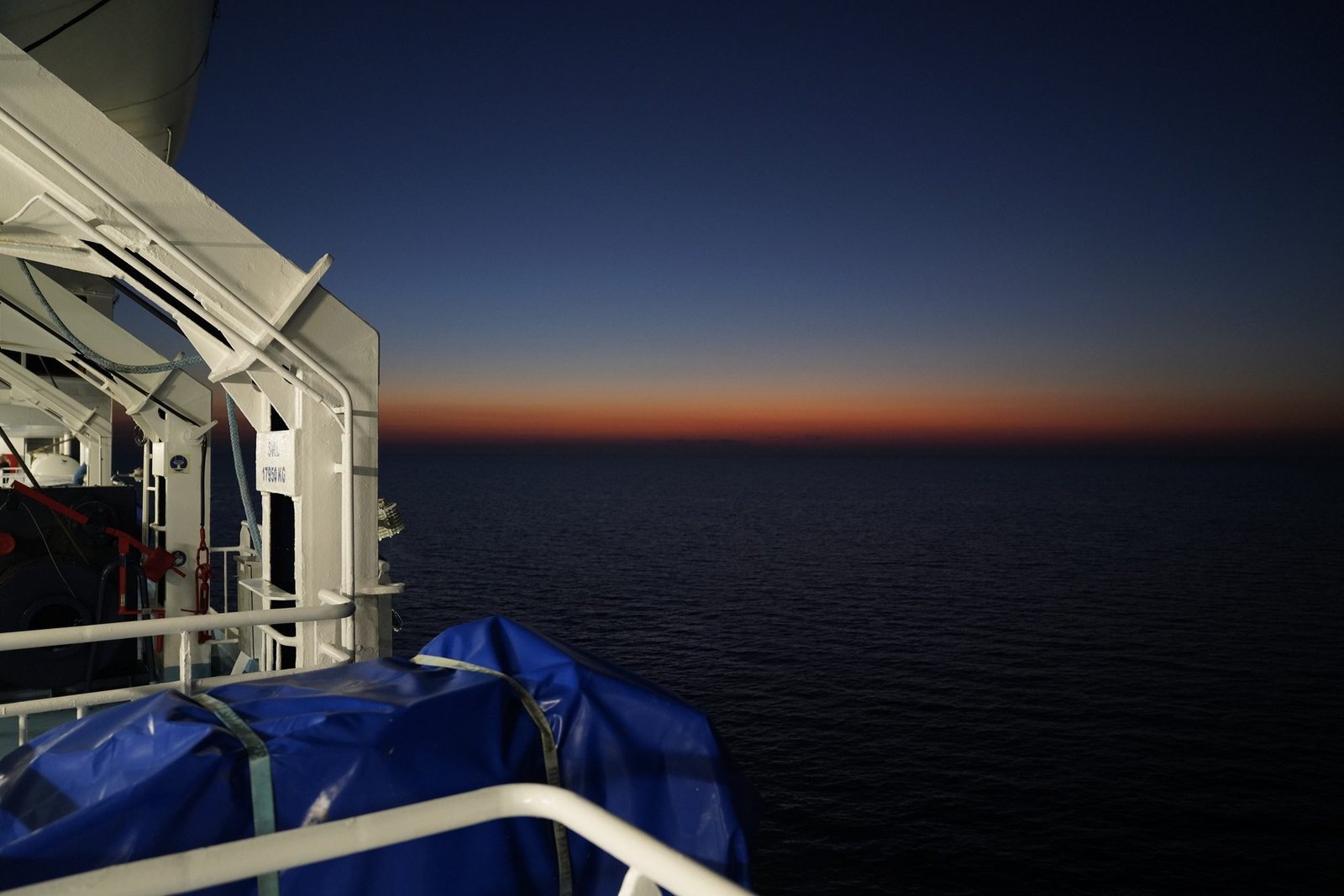 Vue depuis le pont sur une mer sombre, avec une bande orange à l’horizon après le coucher du soleil et un canot de sauvetage à gauche