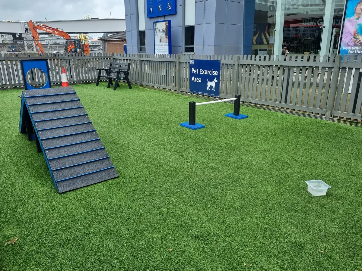 Un espace de promenade en gazon synthétique entouré d’une clôture en bois grise, avec une rampe, une barre de saut, un panneau percé d’un trou rond, un banc, un bac d’eau et le panneau Pet Exercise Area, devant un bâtiment du terminal