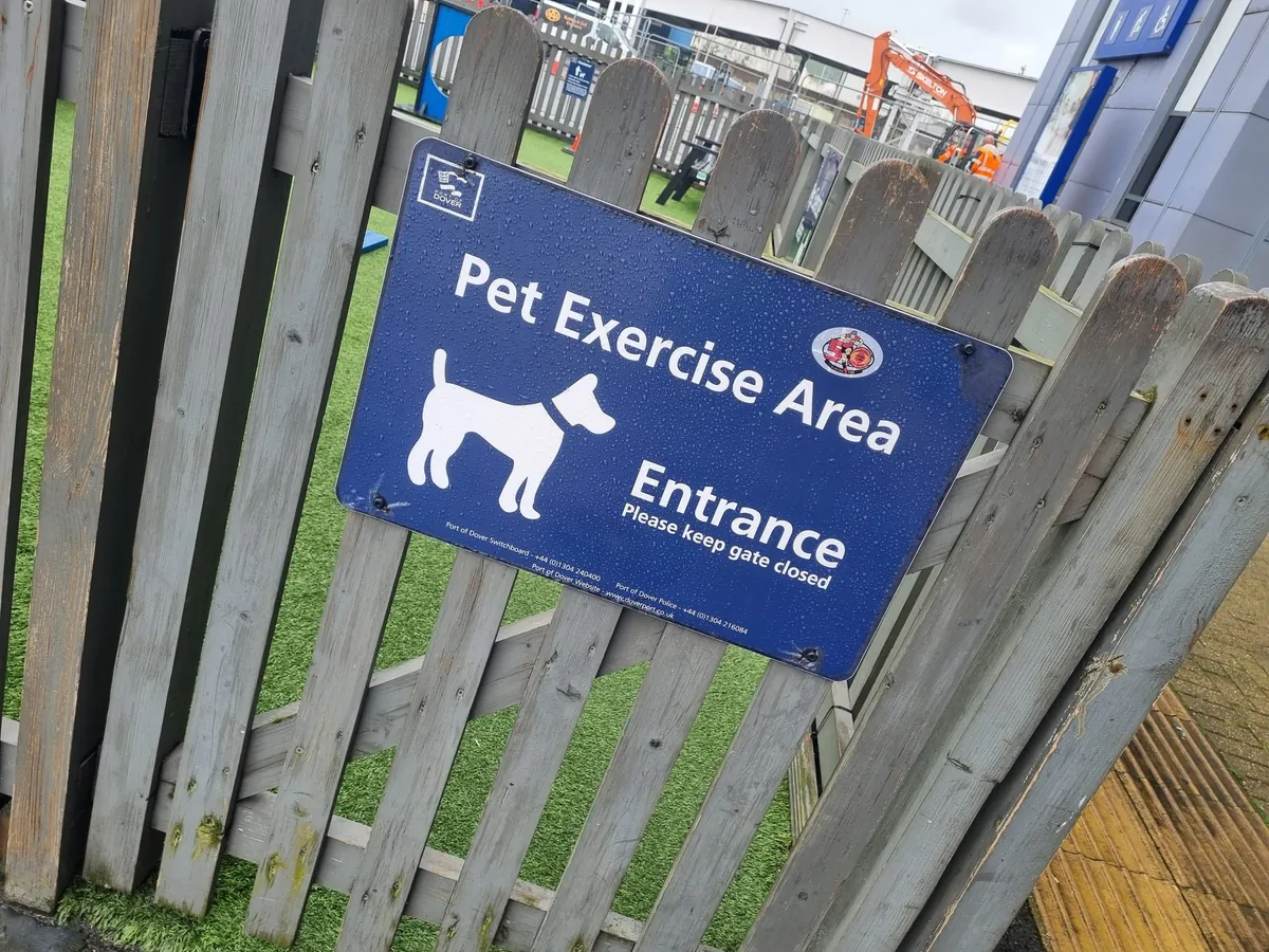 Un panneau bleu du port de Douvres avec Pet Exercise Area, Entrance et Please keep gate closed sur un petit portail en bois gris, avec du gazon synthétique derrière