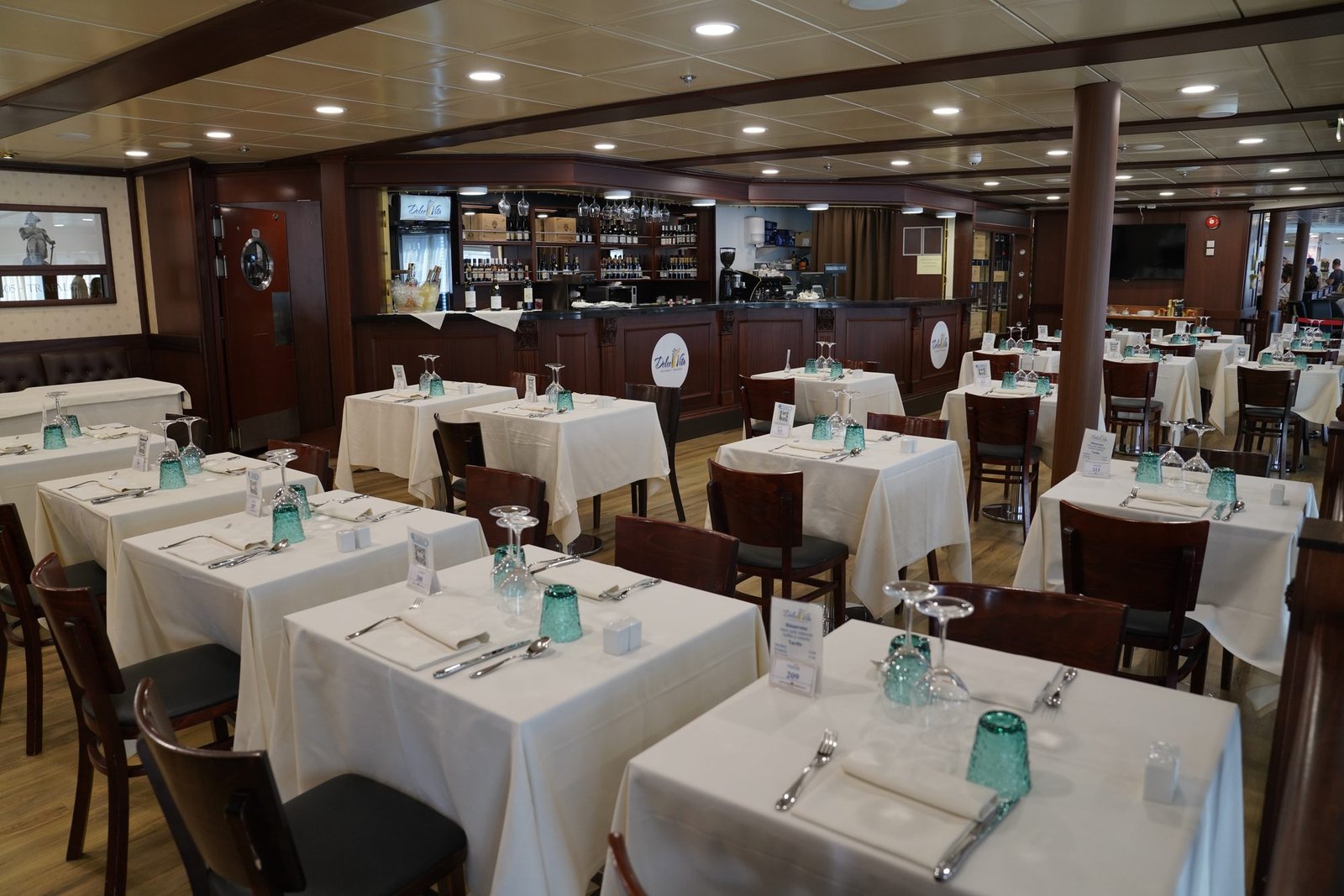 Le restaurant Dolce Vita à bord, avec des tables dressées de nappes blanches et de verres verts, et un bar en bois avec des bouteilles de vin derrière