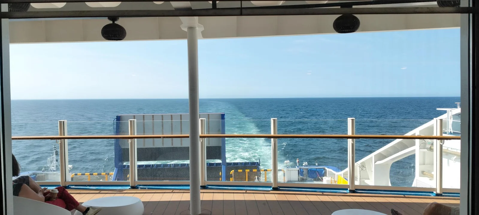 Vue depuis le pont arrière couvert du ferry, par-dessus le bastingage, sur le sillage et la mer, avec un passager dans un fauteuil blanc à gauche