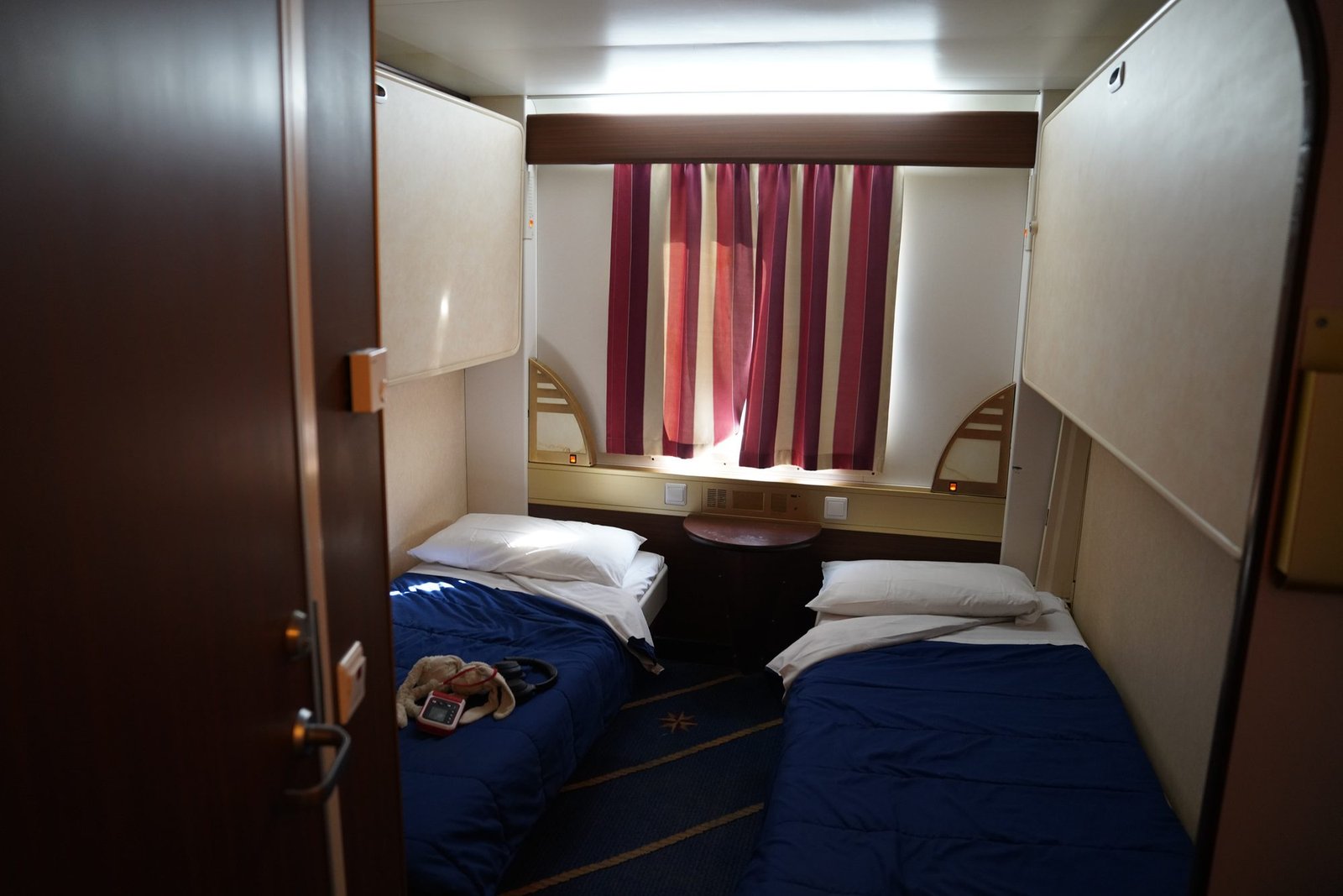 Cabine pour quatre avec deux lits bas aux couvre-lits bleus, deux couchettes rabattues contre les cloisons et un hublot avec un rideau à rayures rouges