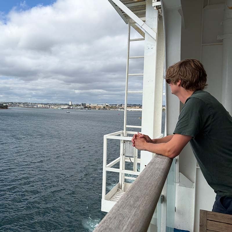 Jochem Vroom en t-shirt vert, accoudé au bastingage en bois d’un ferry, regarde l’eau et une ville côtière sous un ciel nuageux