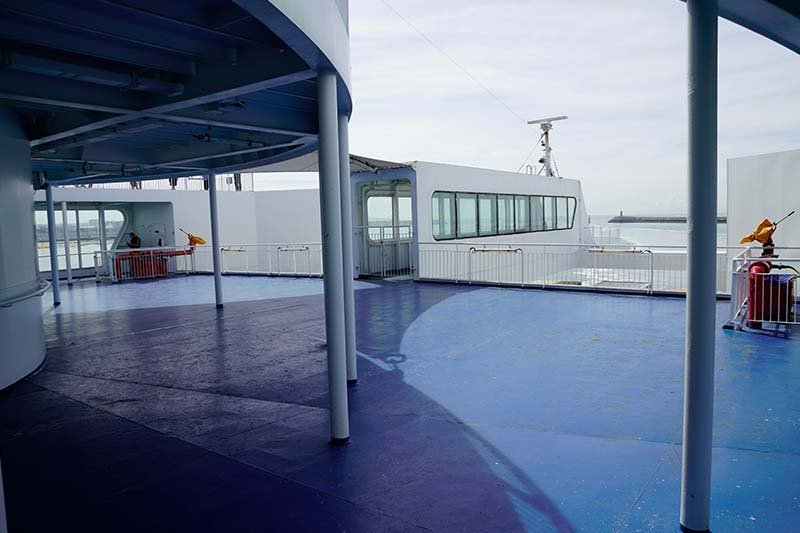 Pont extérieur vide au sol bleu, avec des poteaux et des barrières blancs et une vue sur l’entrée du port, sans un seul banc ni une seule chaise