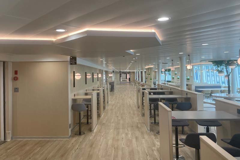 Pont à bord de P&O Ferries avec un plancher en bois clair, des tables hautes et des tabourets de bar dans des niches et des fenêtres sur la droite