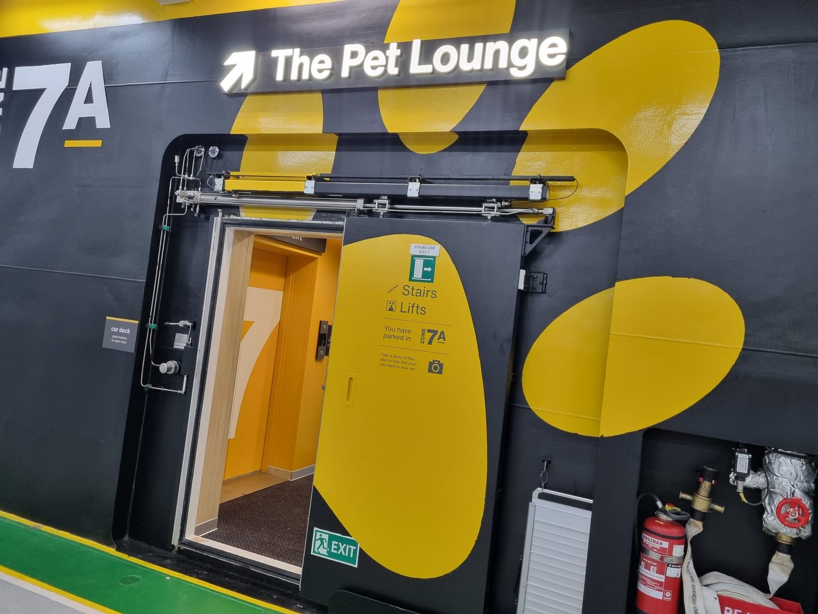Un panneau lumineux The Pet Lounge avec une flèche au-dessus d’une porte vers les escaliers et les ascenseurs, sur un mur noir à grandes taches jaunes, au pont garage près de la zone 7A