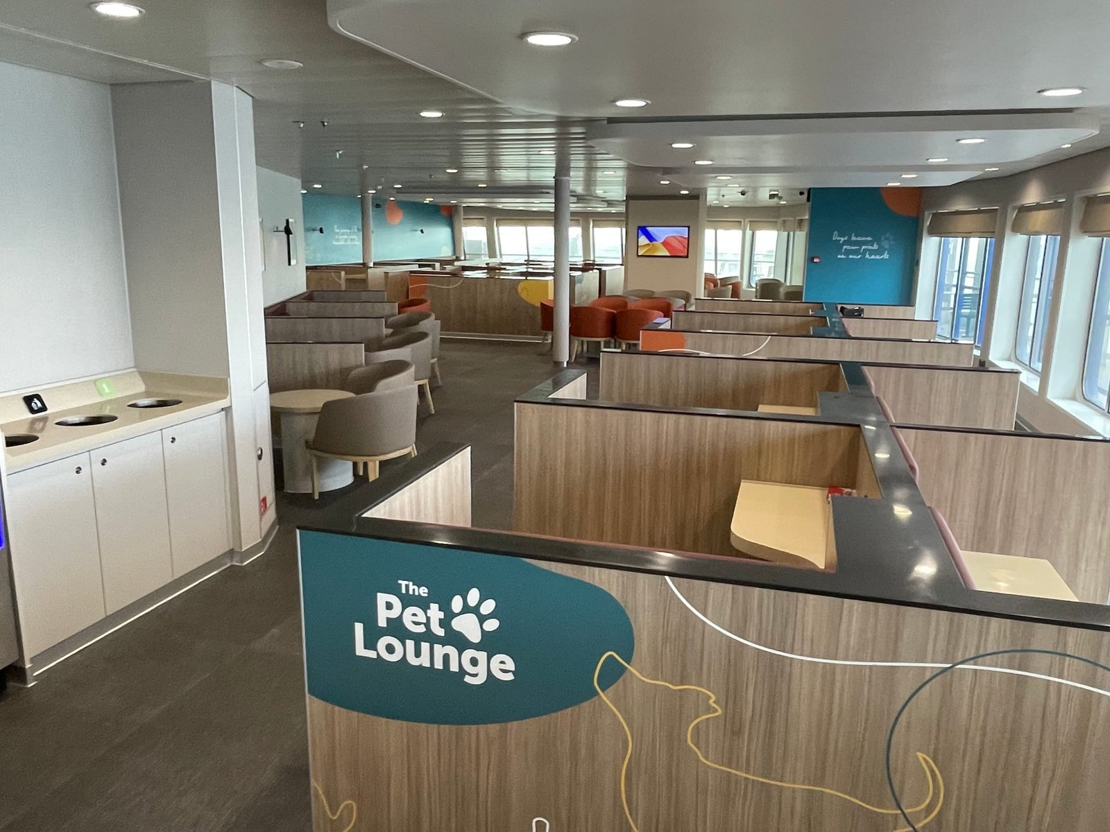 The Pet Lounge à bord : des box séparés par des cloisons en bois, des chaises et des tables rondes, des murs bleu pétrole avec la phrase Dogs leave paw prints on our hearts et des fenêtres sur le côté