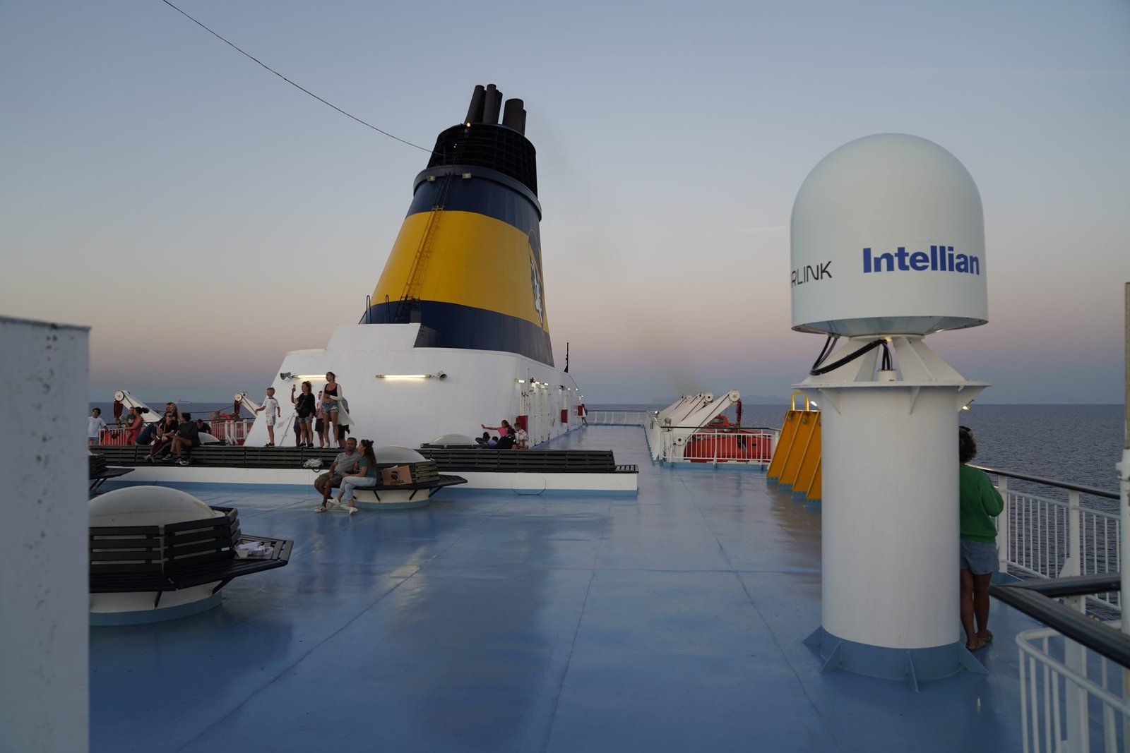 Le pont supérieur du ferry au crépuscule, avec la cheminée jaune, des passagers sur des bancs et un dôme satellite à droite