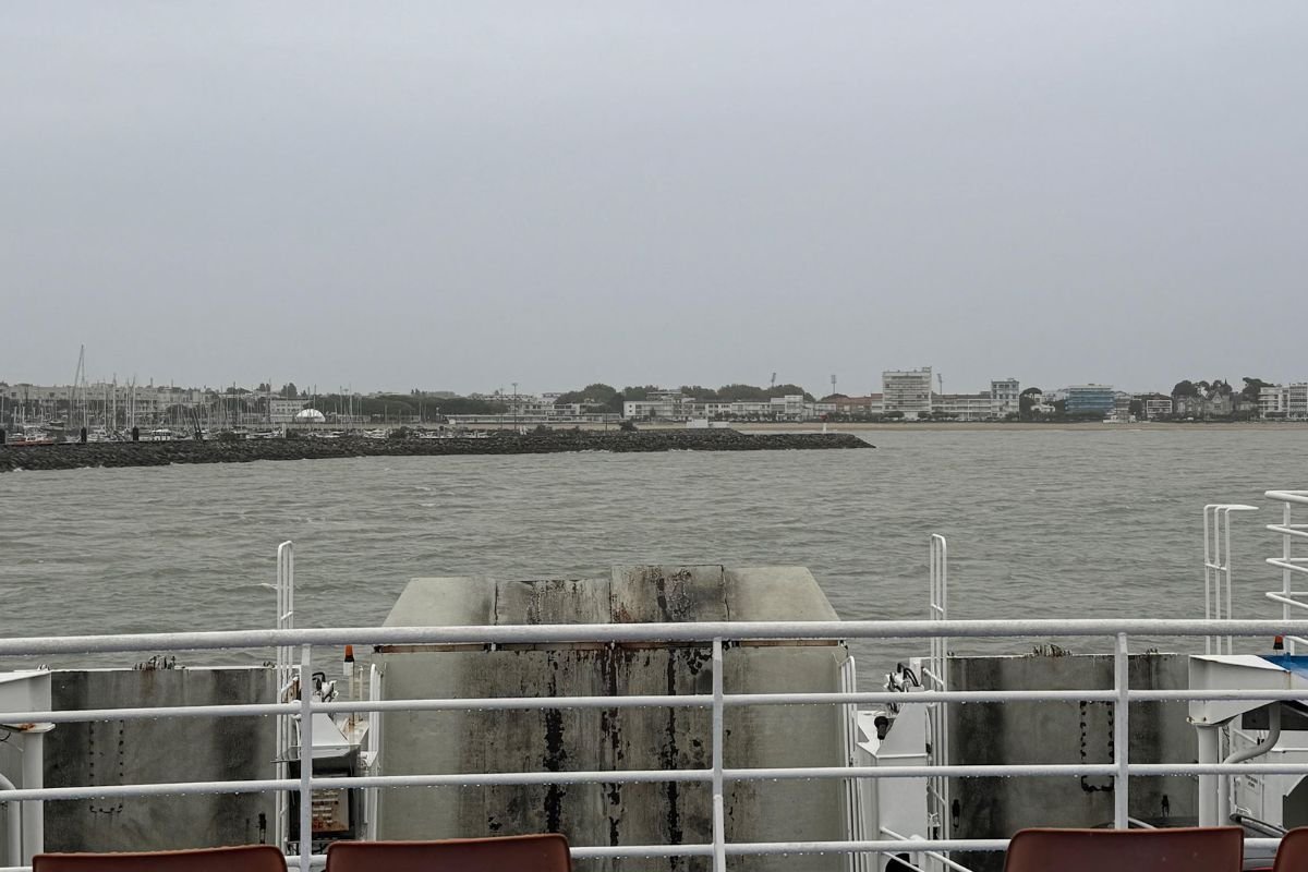 Vue depuis le pont, par-dessus le bastingage et les radeaux de sauvetage, sur une eau grise, avec une digue en pierre, les immeubles et le port de plaisance d’une station balnéaire sur l’autre rive